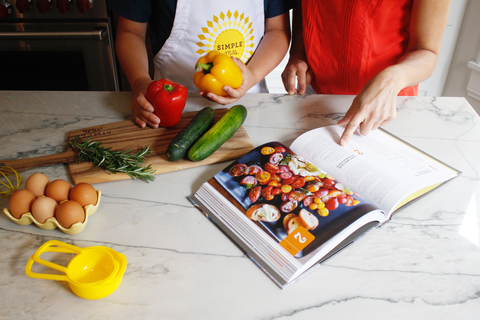 Michelle pointing to a section in cookbook as son holds bell peppers and looks on